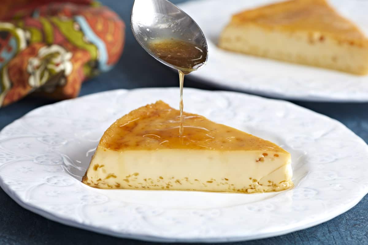 Side view of a slice of flan on a plate with caramel being drizzled over top.
