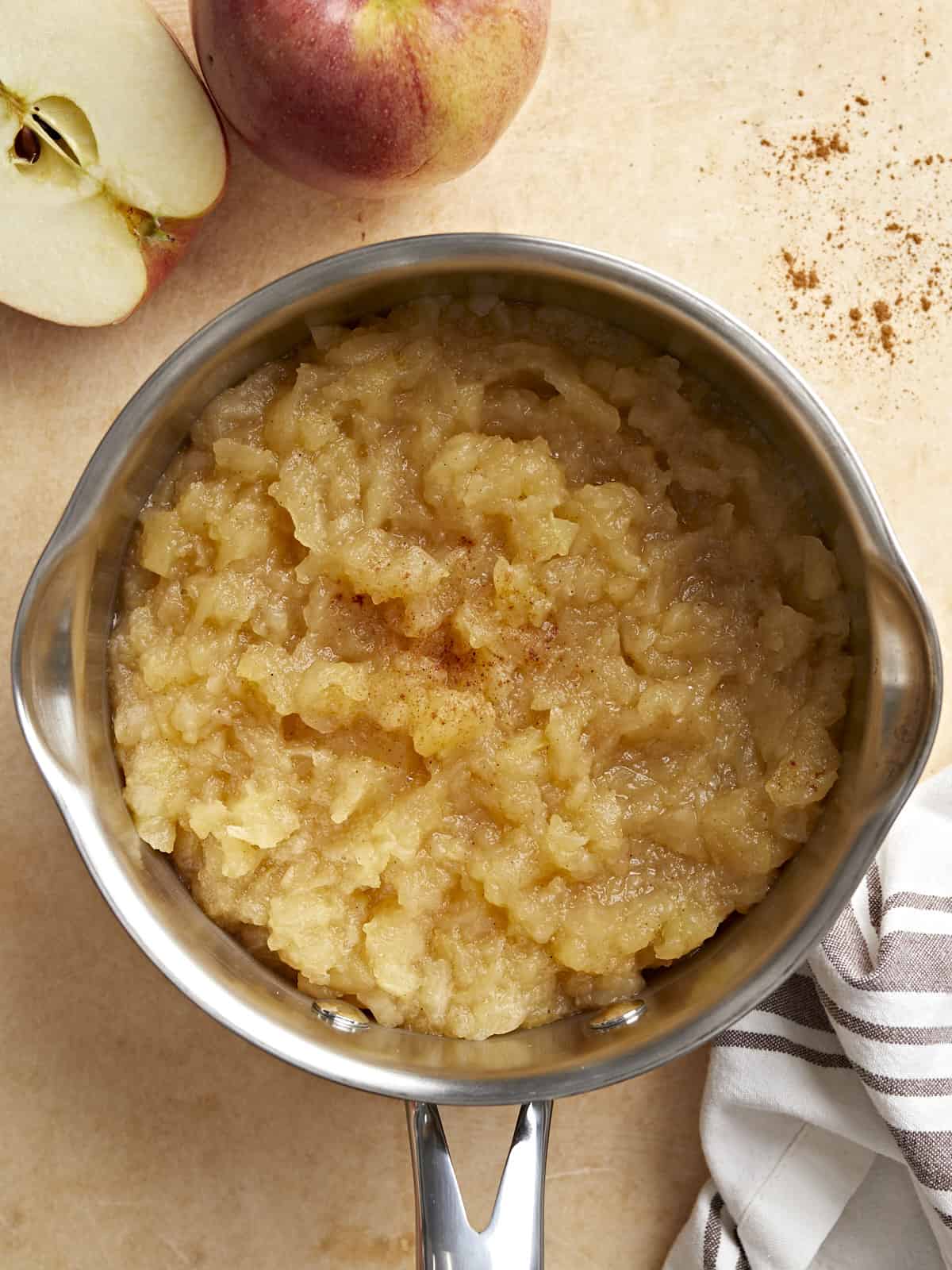 Homemade applesauce in a small saucepan with apples and a napkin on the side.