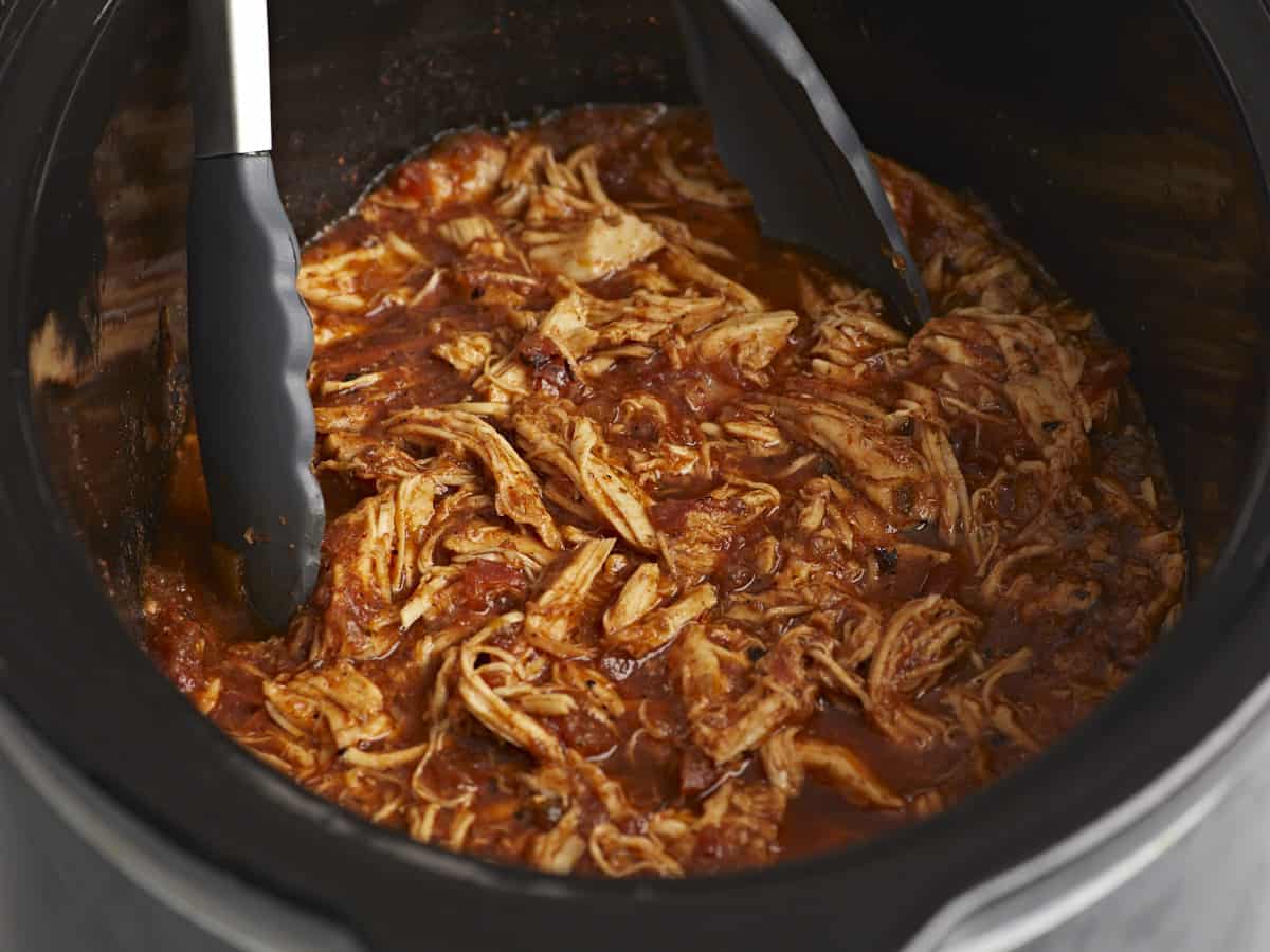 Close up side view of salsa chicken in the crock pot with tongs. 