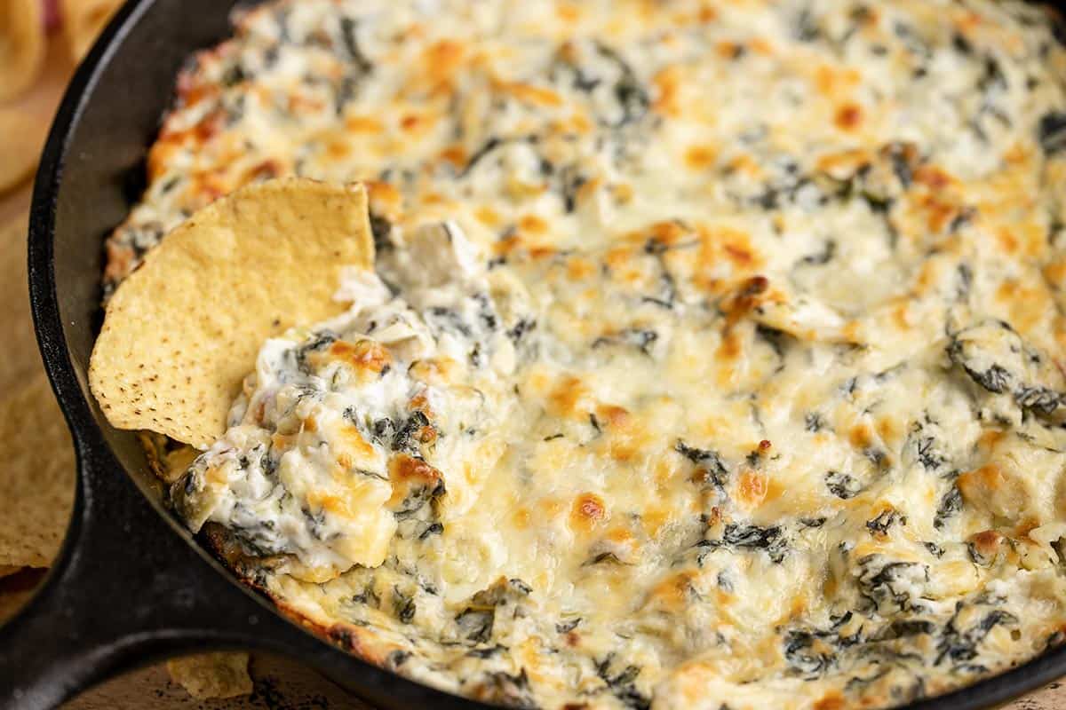 Close up side view of a tortilla chip dipping into the pan of spinach artichoke dip.