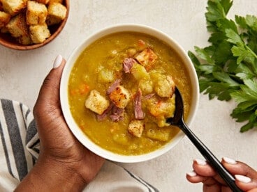 Overhead view of a single bowl of split pea soup with one hand holding the bowl and another hand spooning some of the soup out.