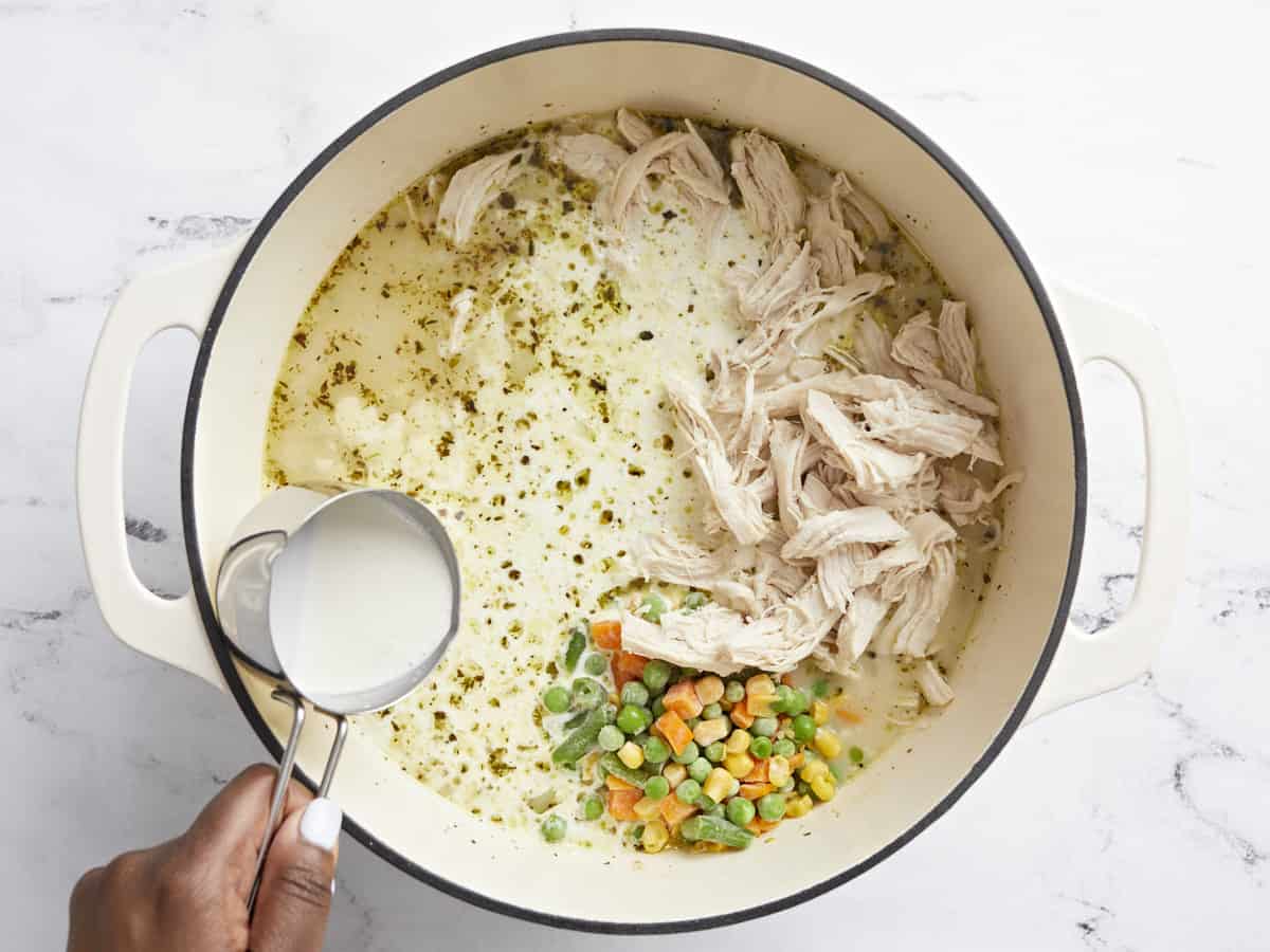 Overhead view of shredded chicken, frozen vegetables, and heavy cream being added to the pot.