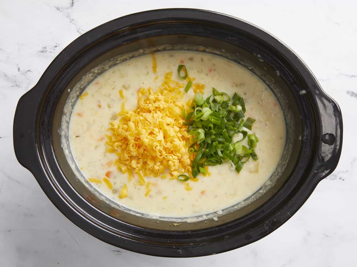 Shredded cheese and green onions being added to potato soup in a slow cooker.
