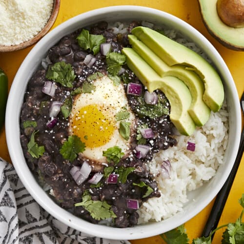 Overhead view of a bowl of black beans with eggs over rice with sliced avocado on the side.