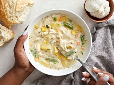 Overhead view of a serving bowl of chicken pot pie soup with one hand holding the bowl and the other hand holding a spoon .