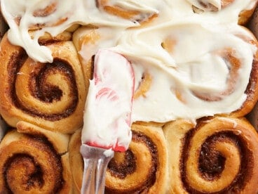 Close up overhead view of cream cheese icing being spread onto homemade cinnamon rolls.