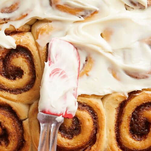 Close up overhead view of cream cheese icing being spread onto homemade cinnamon rolls.