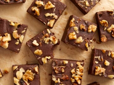 Overhead view of chocolate fudge with walnuts cut into squares on a wooden surface, one with a bite taken out.