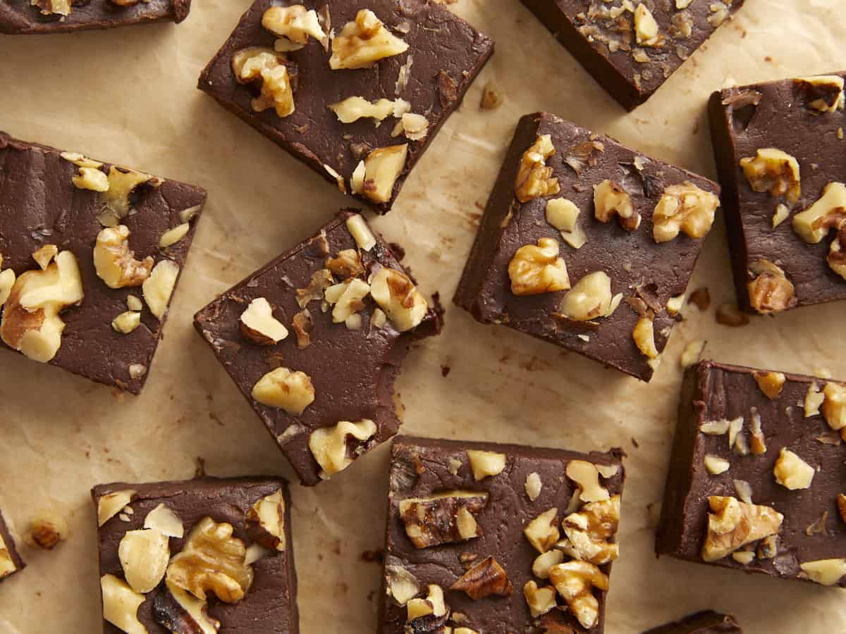 Overhead view of chocolate fudge with walnuts cut into squares on a wooden surface, one with a bite taken out. 