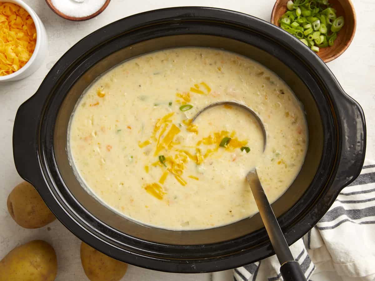 Potato soup in a slow cooker with shredded cheese, sliced green onions and a napkin on the side.