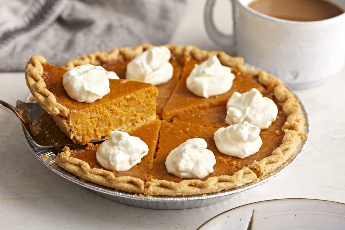 Side view of a whole sweet potato pie with dollops of whipped cream on top and one slice being lifted out of the pie dish.