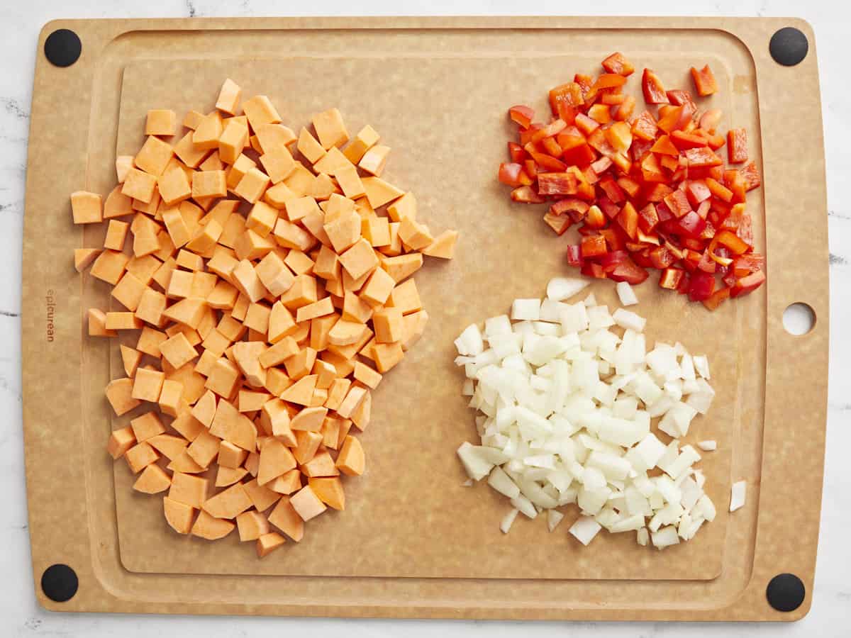 Diced sweet potato, a diced red bell pepper and a diced onion on a cutting board.