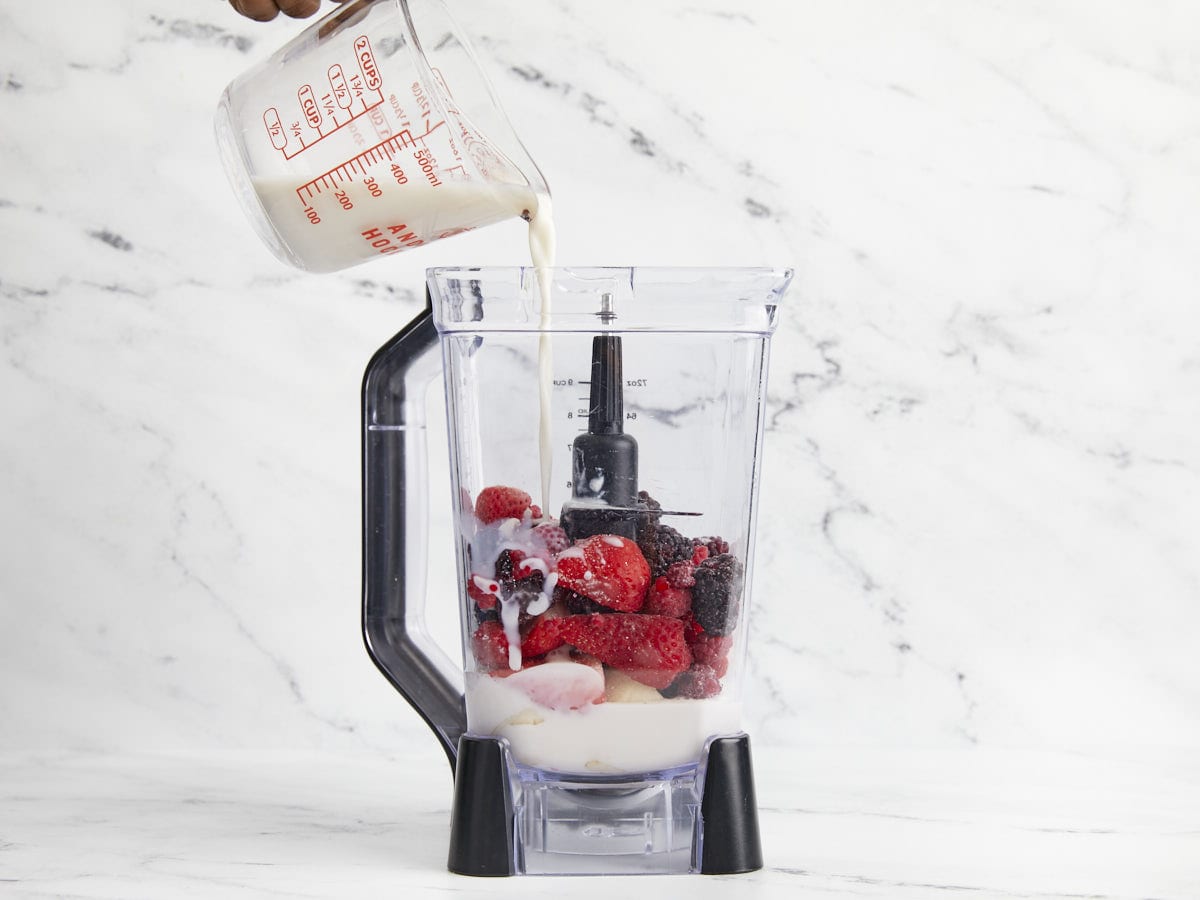 Front view of mixed berry smoothie ingredients in a blender with almond milk being poured inside the blender.