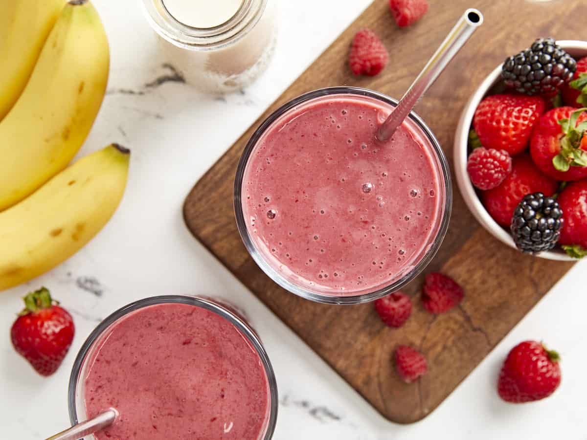 Overhead shot of two mixed berry smoothies in glasses with metal straws and fresh fruit on the side.