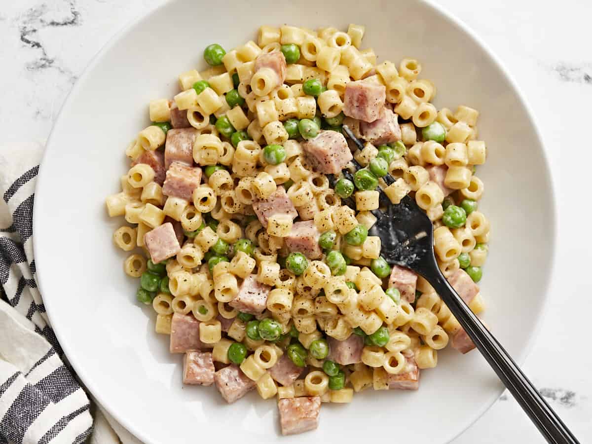 Overhead view of a bowl full of pasta with peas and ham and a fork.