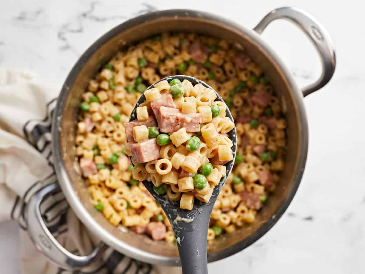 Close up of pasta with ham and peas in a spoon held over the pot. 