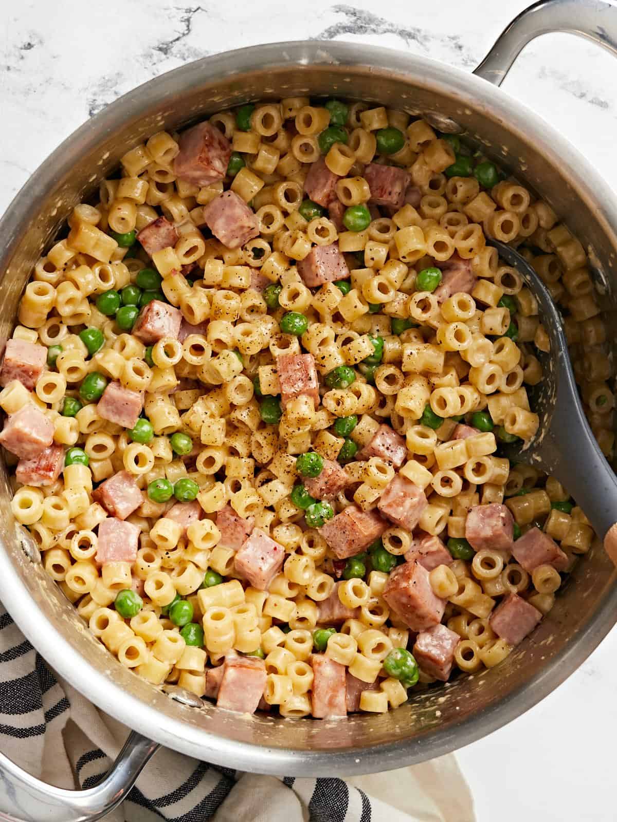 Overhead view of a pot full of pasta with peas and ham, a spoon in the side.