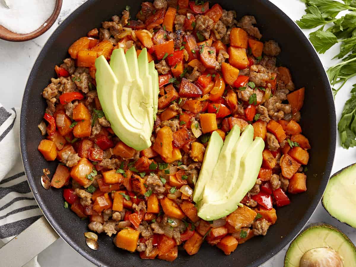 Overhead view of sweet potato hash in a skillet garnished with sliced avocado.