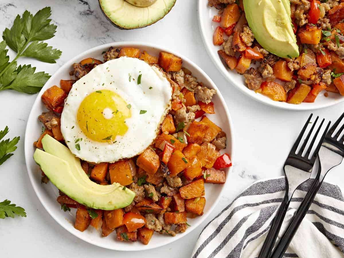 Overhead view of sweet potato hash on two plates topped with sliced avocado and a fried egg.