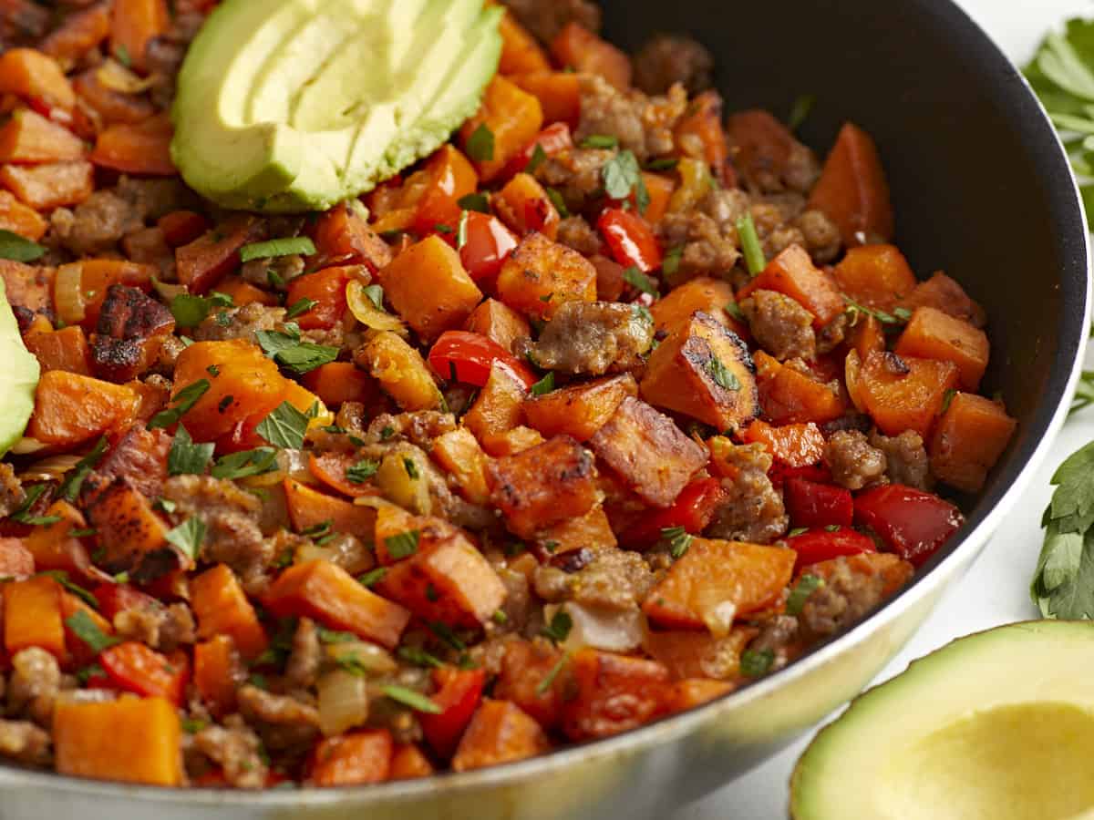 Side view of sweet potato hash in a skillet with sliced avocado on top.