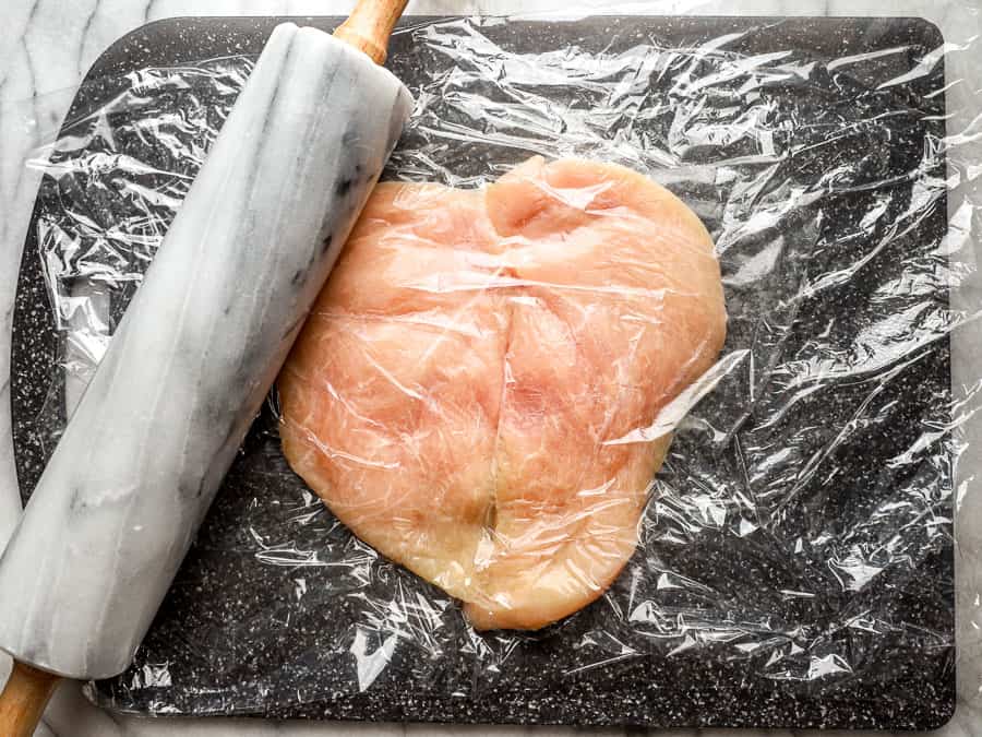 a butterflied chicken breast under plastic wrap with a rolling pin.