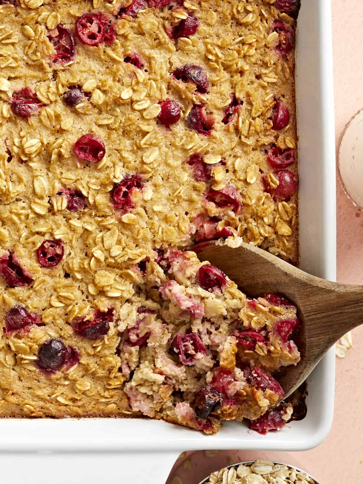 Close up overhead view of a casserole dish full of cranberry apple baked oatmeal and a wooden spoon scooping some out.