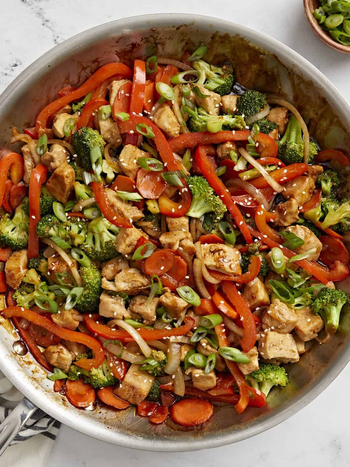 Overhead view of chicken stir fry in a large skillet with sliced green onions on the side.