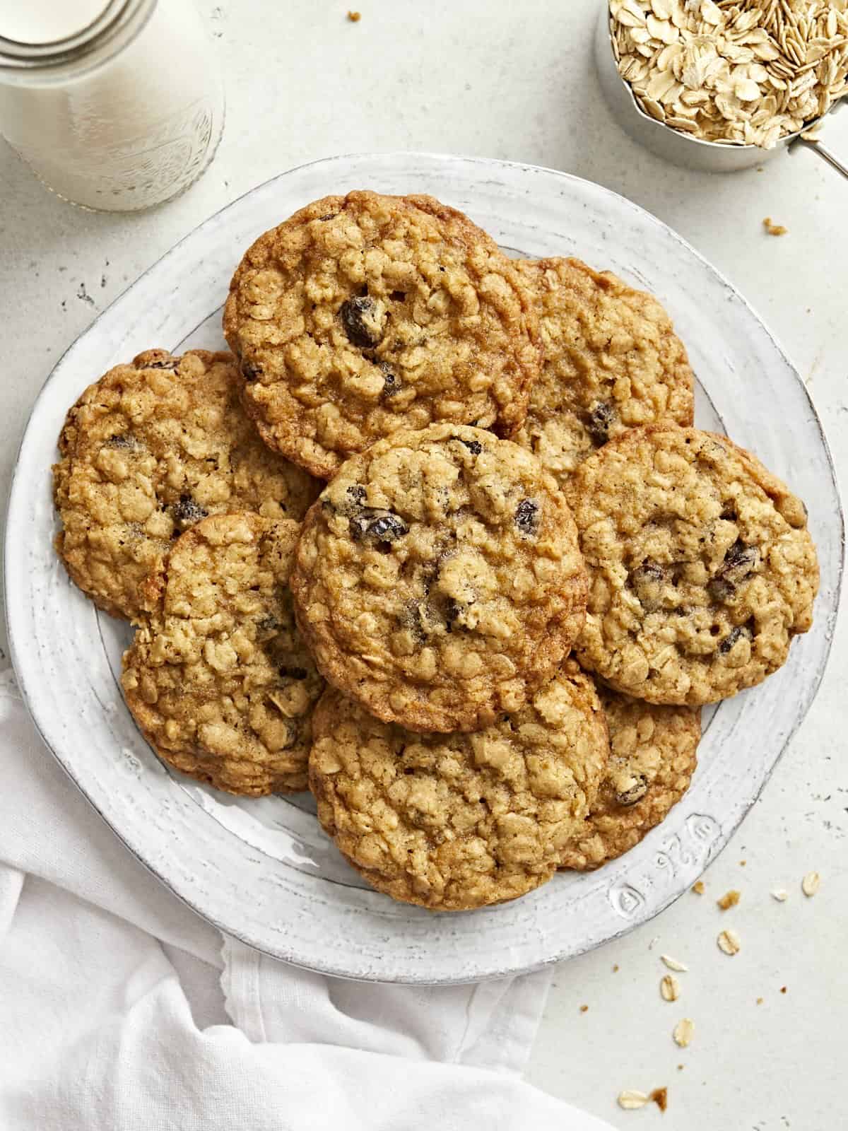 Overhead view of a pile of oatmeal cookies on a plate with a white napkin and a cup of milk on the side.