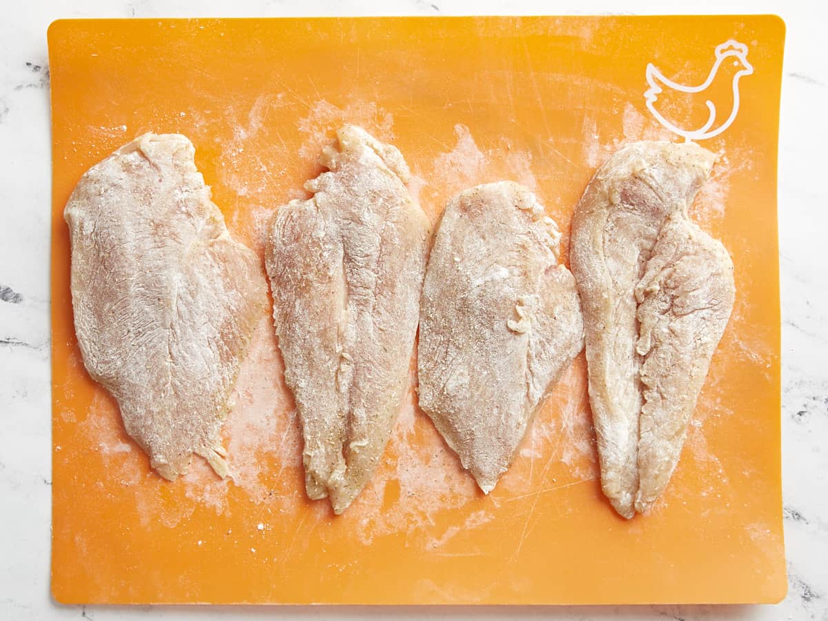 Four chicken breast cutlets, seasoned, and floured on a cutting board.
