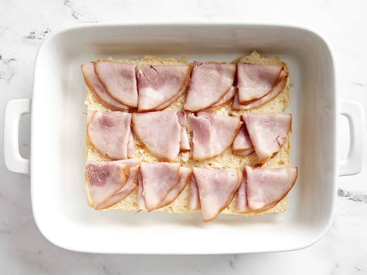 12 ham slices added on top of hawaiian rolls in a white baking dish.