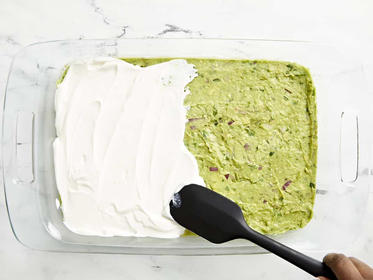Sour cream being spread on top of guacamole with a black spatula.