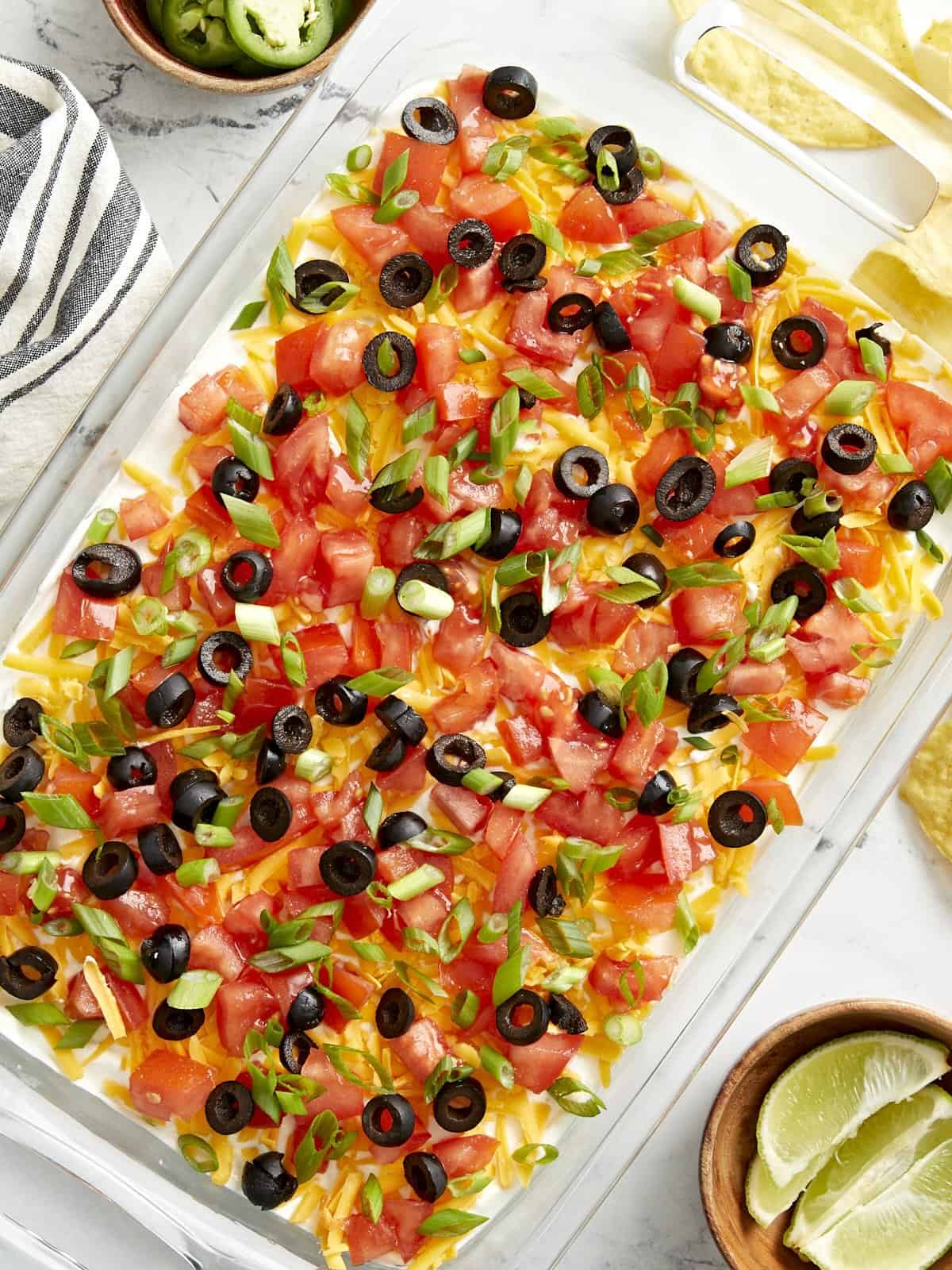 Overhead view of 7 layer dip in a glass casserole dish with a napkin, chips, and lime wedges on the side.