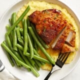 overhead view of a partially sliced air fryer pork chop on a white plate with grits and green beans.