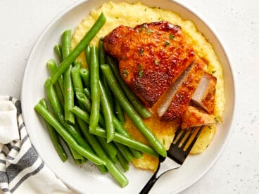 overhead view of a partially sliced air fryer pork chop on a white plate with grits and green beans.