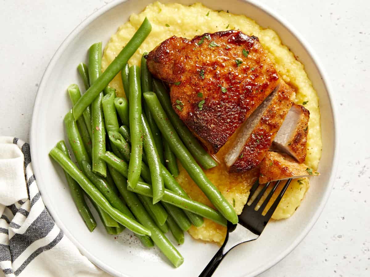 overhead view of a partially sliced air fryer pork chop on a white plate with potatoes and green beans.