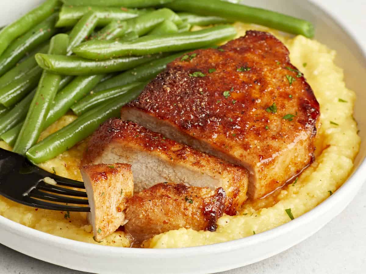 three-quarters view of a partially sliced air fryer pork chop on a white plate with grits and green beans.