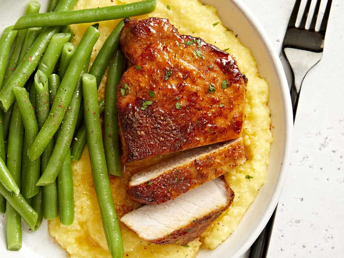 partial overhead view of a partially sliced air fryer pork chop on a white plate with grits and green beans.