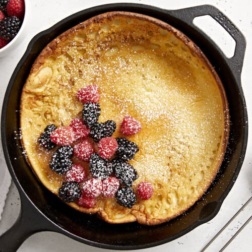 Overhead view of a dutch baby in a cast iron skillet with fresh berries in the center and maple syrup on the side.