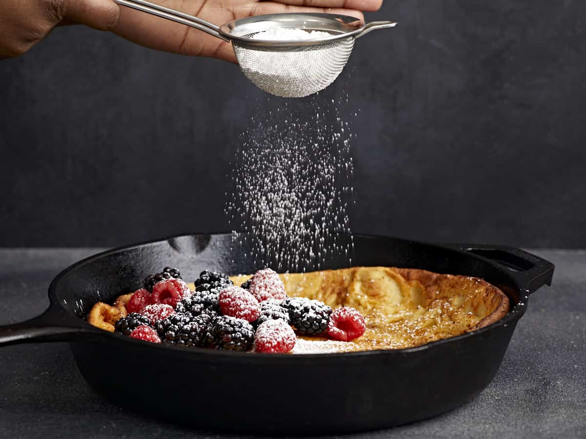 Side view of a Dutch baby in a cast iron skillet with powdered sugar being dusted over the top.