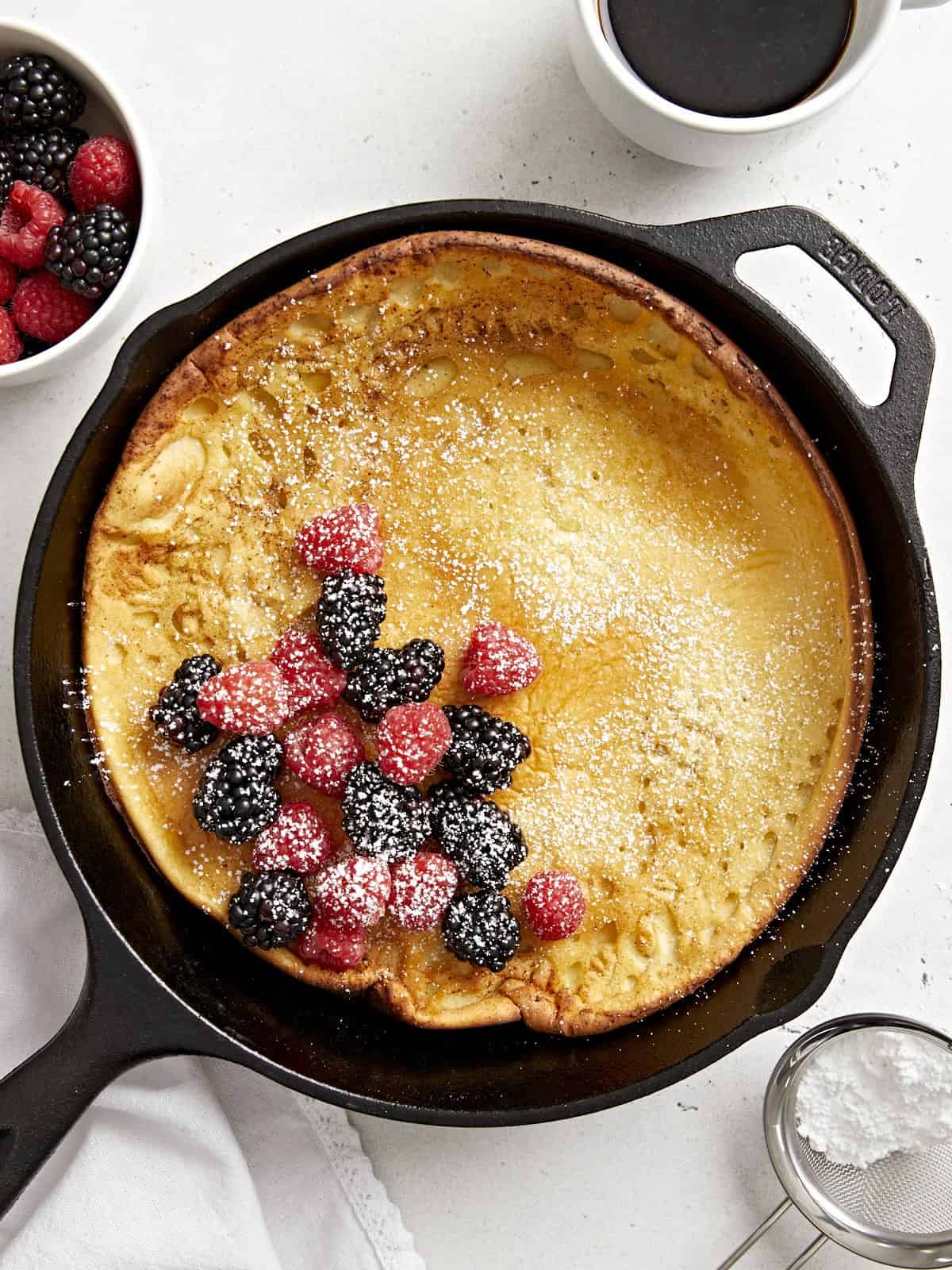 Overhead view of a dutch baby in a cast iron skillet with fresh berries in the center and maple syrup on the side.