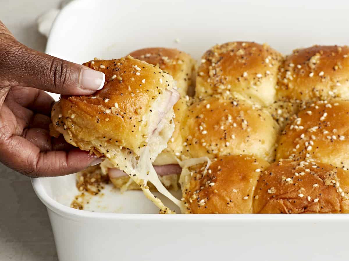 Front side view of a hand pulling one ham and cheese slider out of a casserole baking dish.