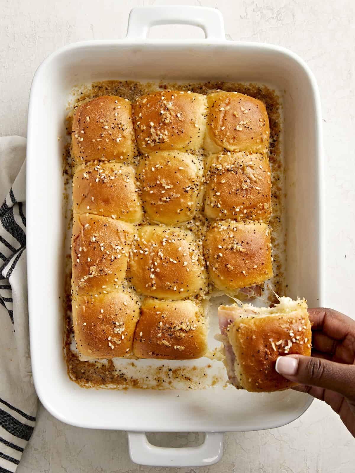 Overhead view of ham and cheese sliders in a baking dish with a hand pulling one slider out of the dish.