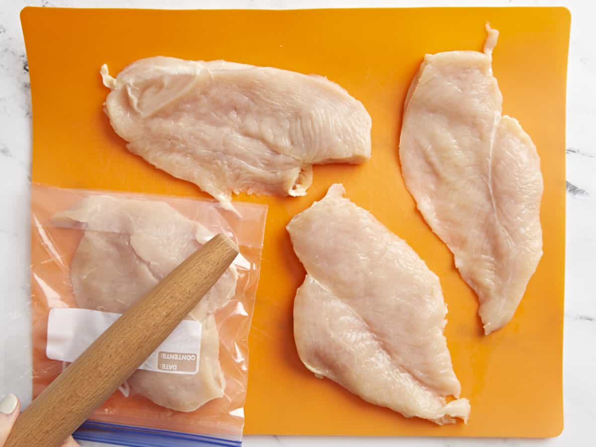 A rolling pin pounding chicken breasts to even thickness on an orange cutting board.