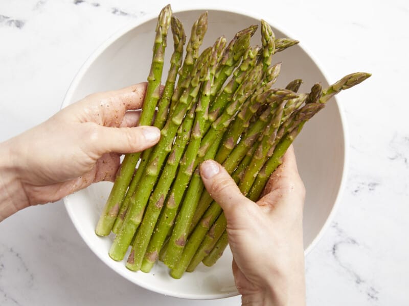 hands tossing asparagus in a white bowl.