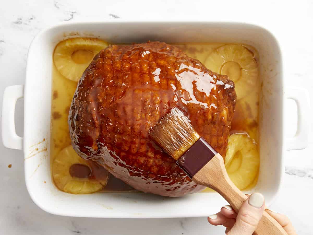 Spread glaze on a baked ham in a white baking dish with pineapple rings.