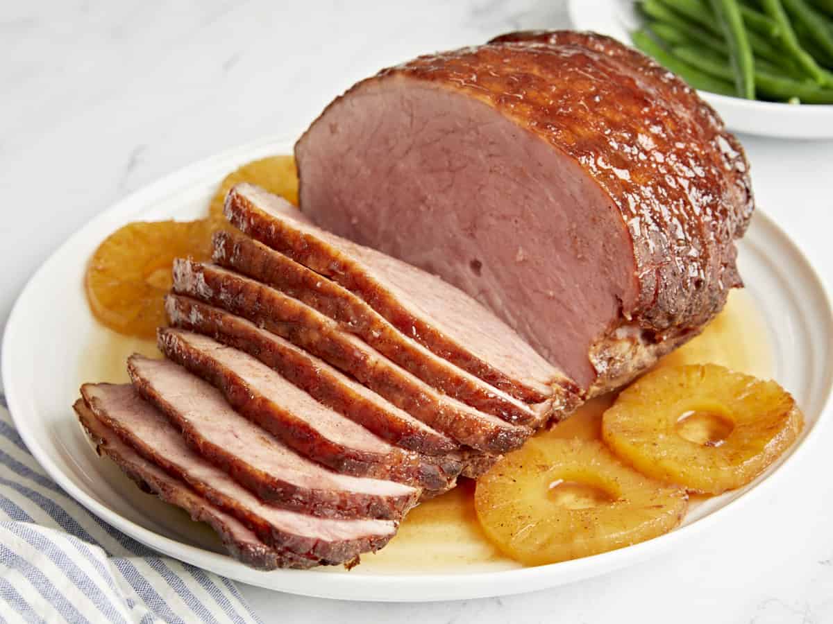 Three-quarter view of a partially sliced ​​baked ham on a white plate with pineapple rings.
