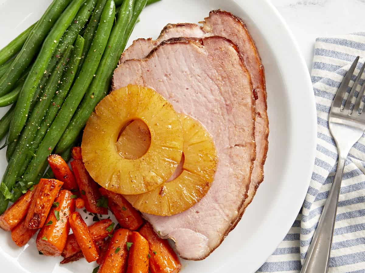 overhead view of slices of baked ham topped with pineapple rings on a white plate with carrots and green beans.
