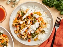 overhead view of buffalo beans and greens over white rice on a white plate with white sauce drizzled over top.