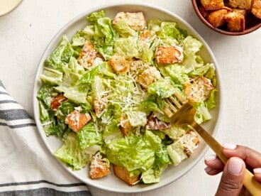 Overhead view of a plate of Caesar salad with a hand holding a fork eating a bite on the side.