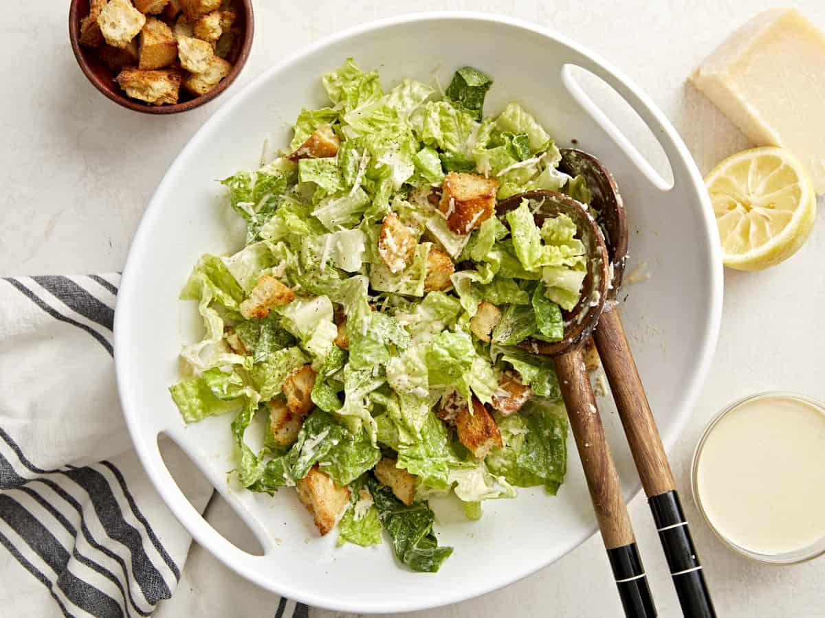 Overhead view of a serving bowl of caesar salad with a napkin, croutons, caesar dressing, and parmesan cheese on the side.